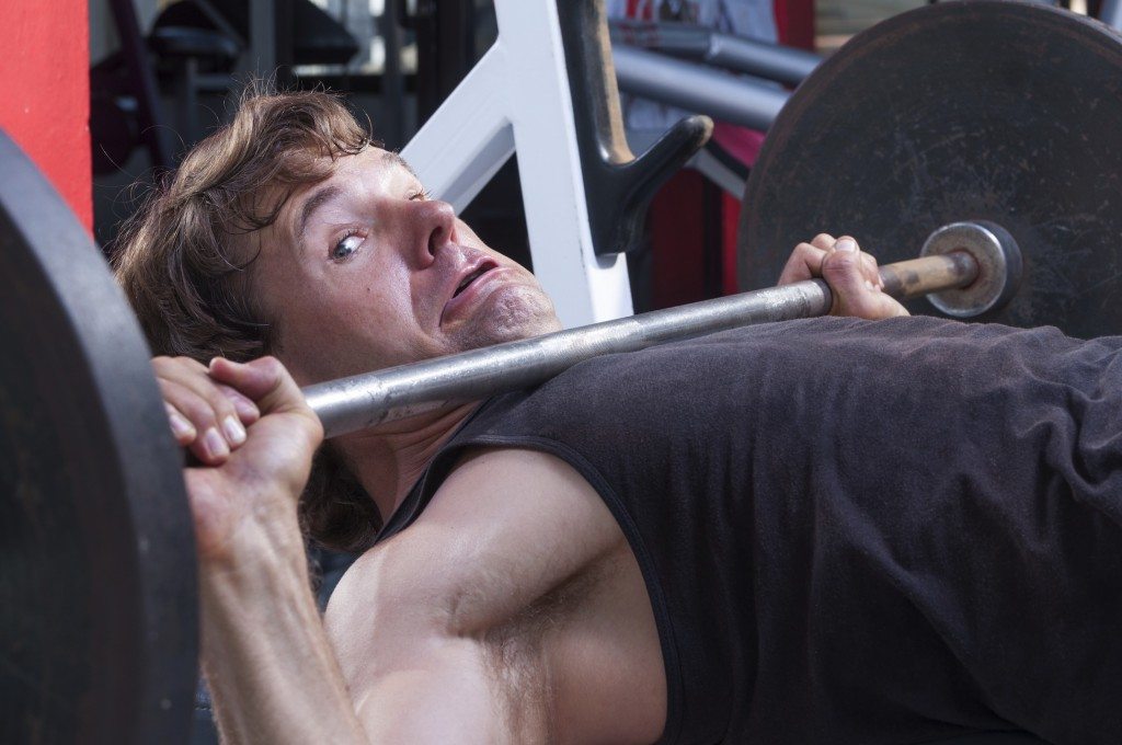 Caucasian man working out in gym is stuck under heavy barbell on bench press