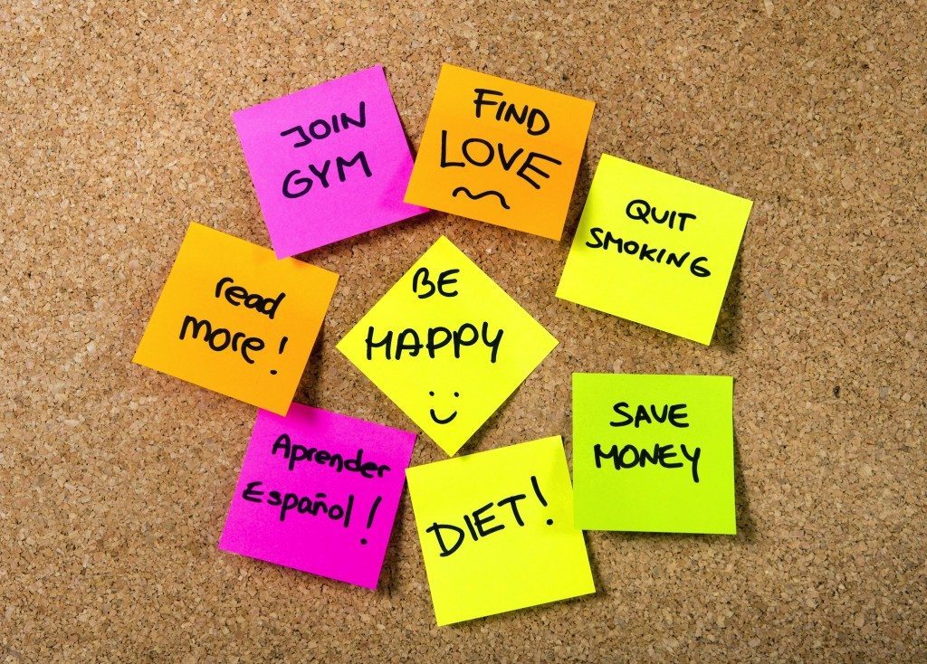 Group of New year Resolutions Post it Notes on pink, yellow, orange and green on cork board written with message of diet, join gym, find love, quit smoking and be happy